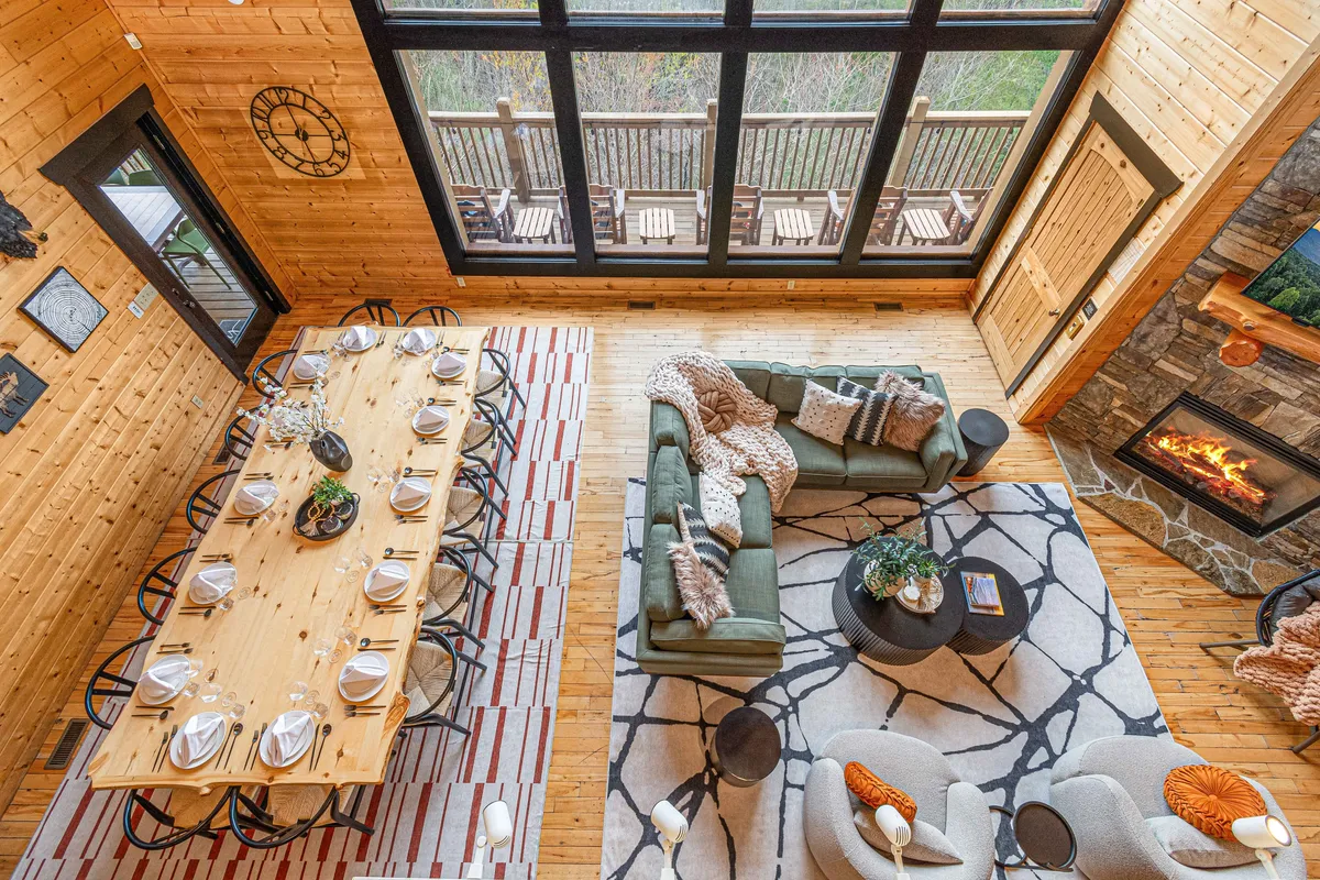 A bright, open-plan interior featuring a spacious dining table set for a group, a comfortable seating area nearby, a stone fireplace, and expansive windows letting in natural light and scenic views of nature. The overall ambiance is welcoming and designed for socializing, meals, and relaxation in a central gathering space.