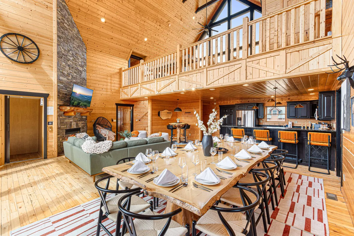 A wide-angle view of an expansive wooden dining table surrounded by chairs, adjacent to a kitchen island and a cozy sofa with decorative pillows. The stone feature wall and large windows add rustic charm and bring in natural light, while the setting invites large gatherings and intimate conversations alike.