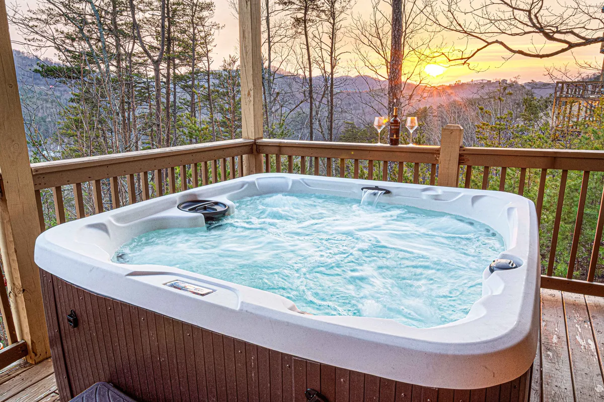 An inviting outdoor scene featuring a bubbling hot tub on a wooden deck at sunset, with champagne glasses nearby suggesting celebration and group enjoyment. The mountain view completes the relaxing, social atmosphere.