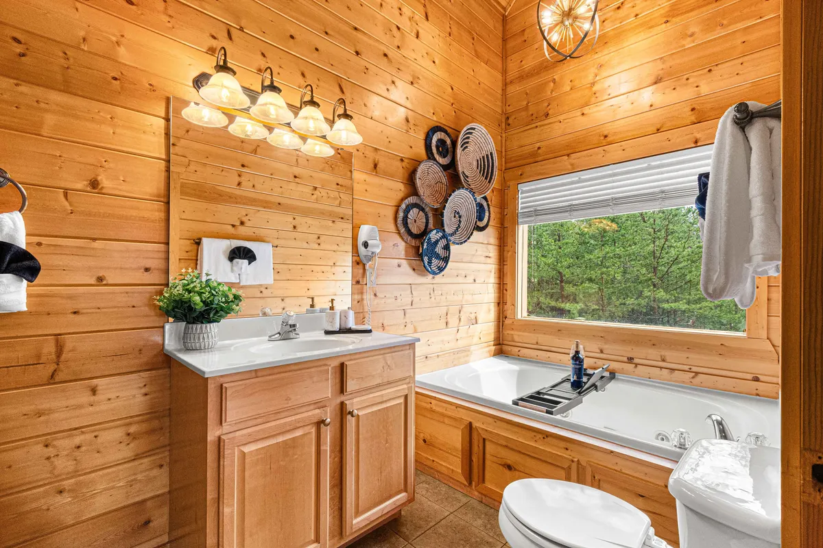 A luxurious bathroom with wooden walls, a freestanding bathtub beside a large window, elegant light fixtures, and a vanity area. The ambiance is serene and refreshing, perfect for relaxing in a private setting with natural views.