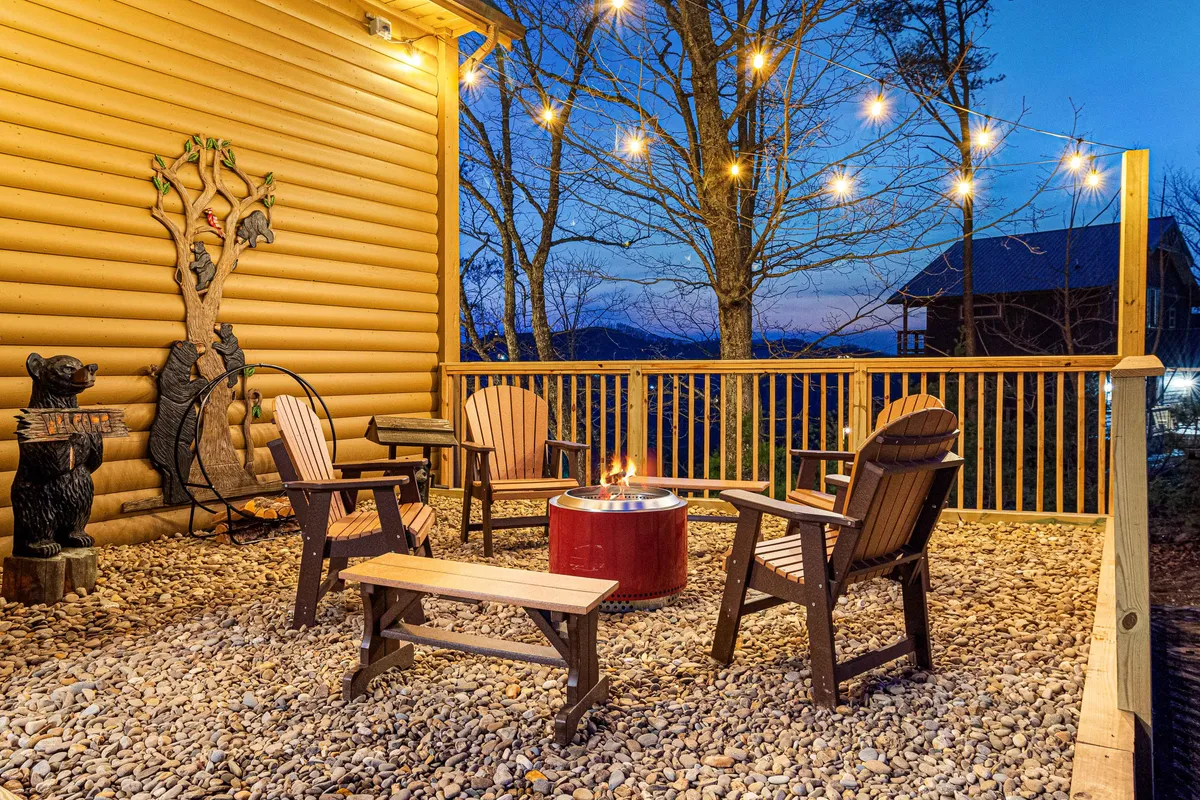 A cozy outdoor fire pit area surrounded by wooden chairs and string lights, with a whimsical bear sculpture adding character. The setting is nestled within lush greenery, inviting guests to gather, unwind, and enjoy the mountain air—perfect for both people and pets.