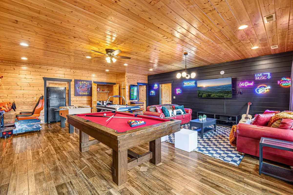A lively game room featuring a pool table, red sofas, arcade game machines, neon signs, and a television. The space is well-lit and designed for group activities and entertainment, inviting guests to enjoy friendly competition and social gatherings.