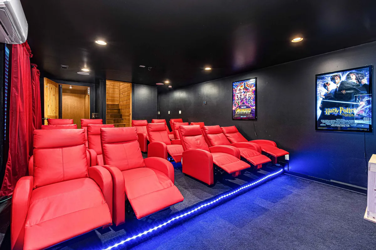 A dedicated theater room with plush red reclining chairs, movie posters on the walls, and ambient lighting. The setup is intimate and immersive, capturing the excitement of a home cinema experience and evoking the energy of movie nights or karaoke sessions.