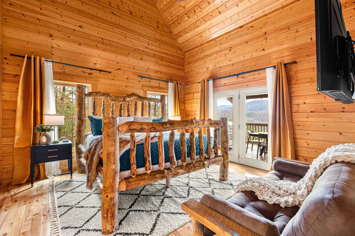 This image shows a spacious, serene bedroom featuring a rustic log bed, large windows flooding the room with natural light, an inviting armchair, and a decorative rug. The design emphasizes comfort and privacy, with views of the surrounding mountains visible from the bed.