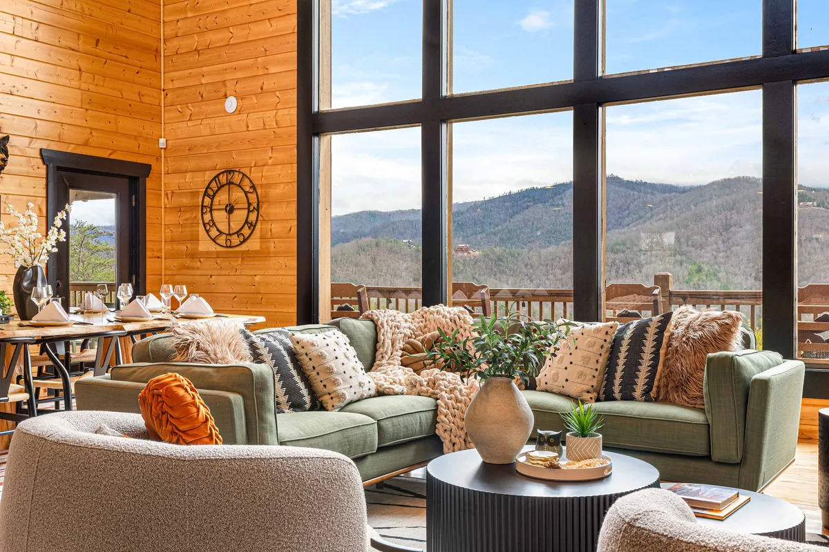 A cozy interior living space featuring large windows that frame a beautiful mountain view, a plush green sofa with decorative pillows, and a dining table elegantly set for guests. Throws and pillows add warmth, while the ambiance suggests both a gathering space for meals and relaxation with family or friends.